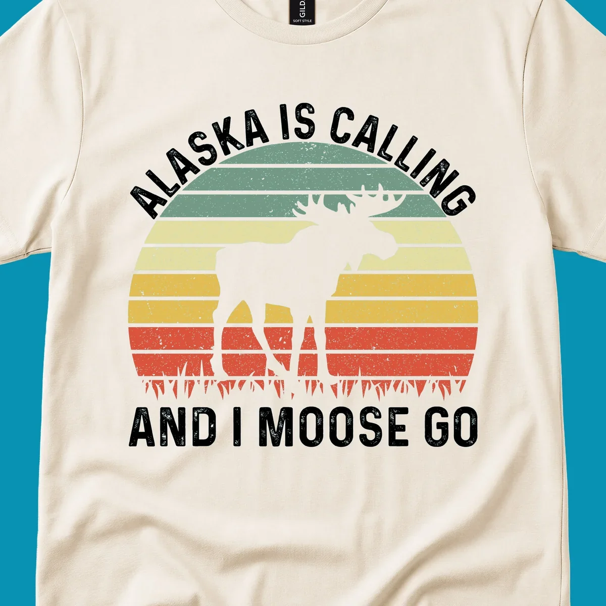 Distressed Alaska calling moose go design with moose and fish silhouettes against striped landscape layers in teal, yellow, and red