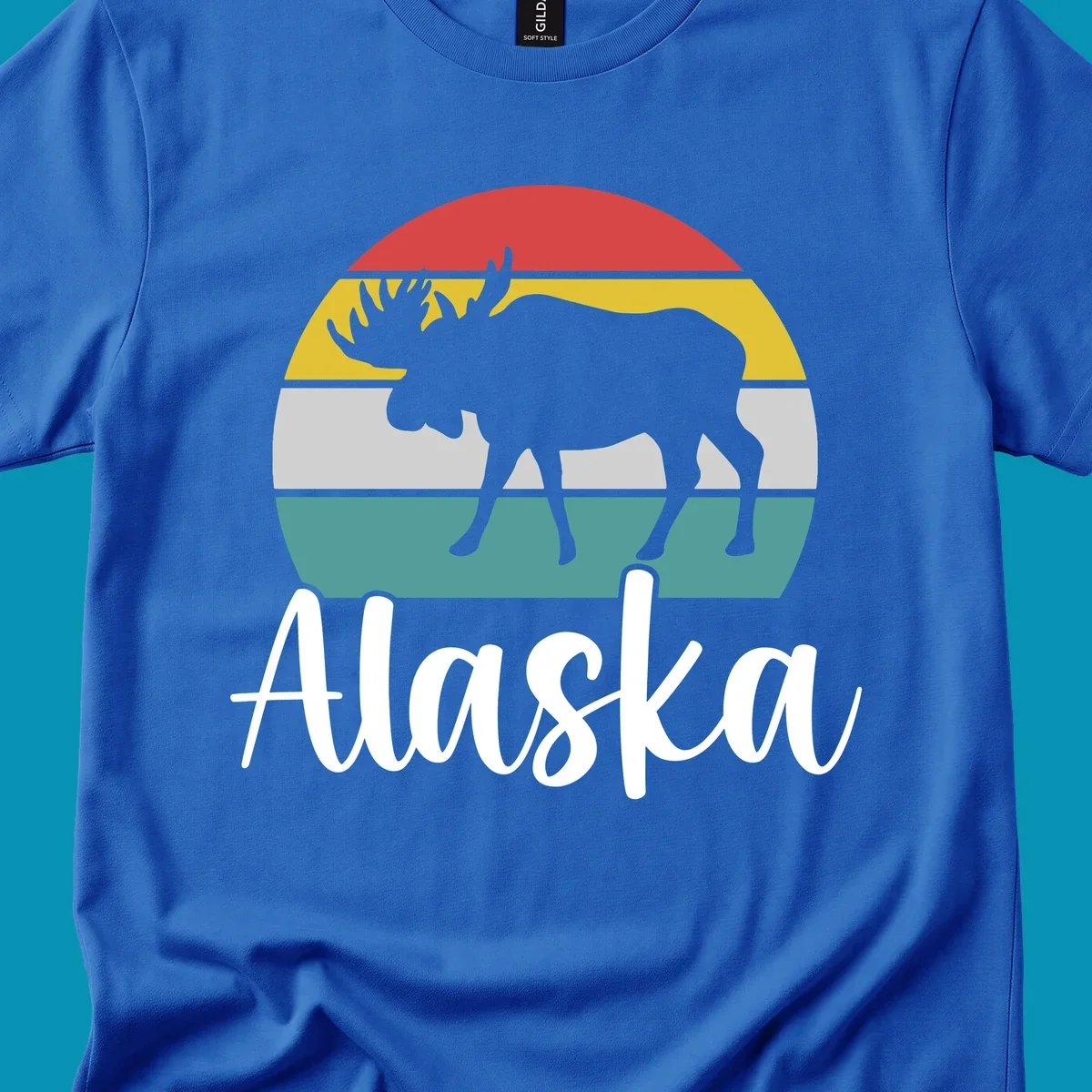 White moose silhouette against red, gold, gray, and teal horizontal stripes in a circular badge design representing Alaska mountains and wildlife.