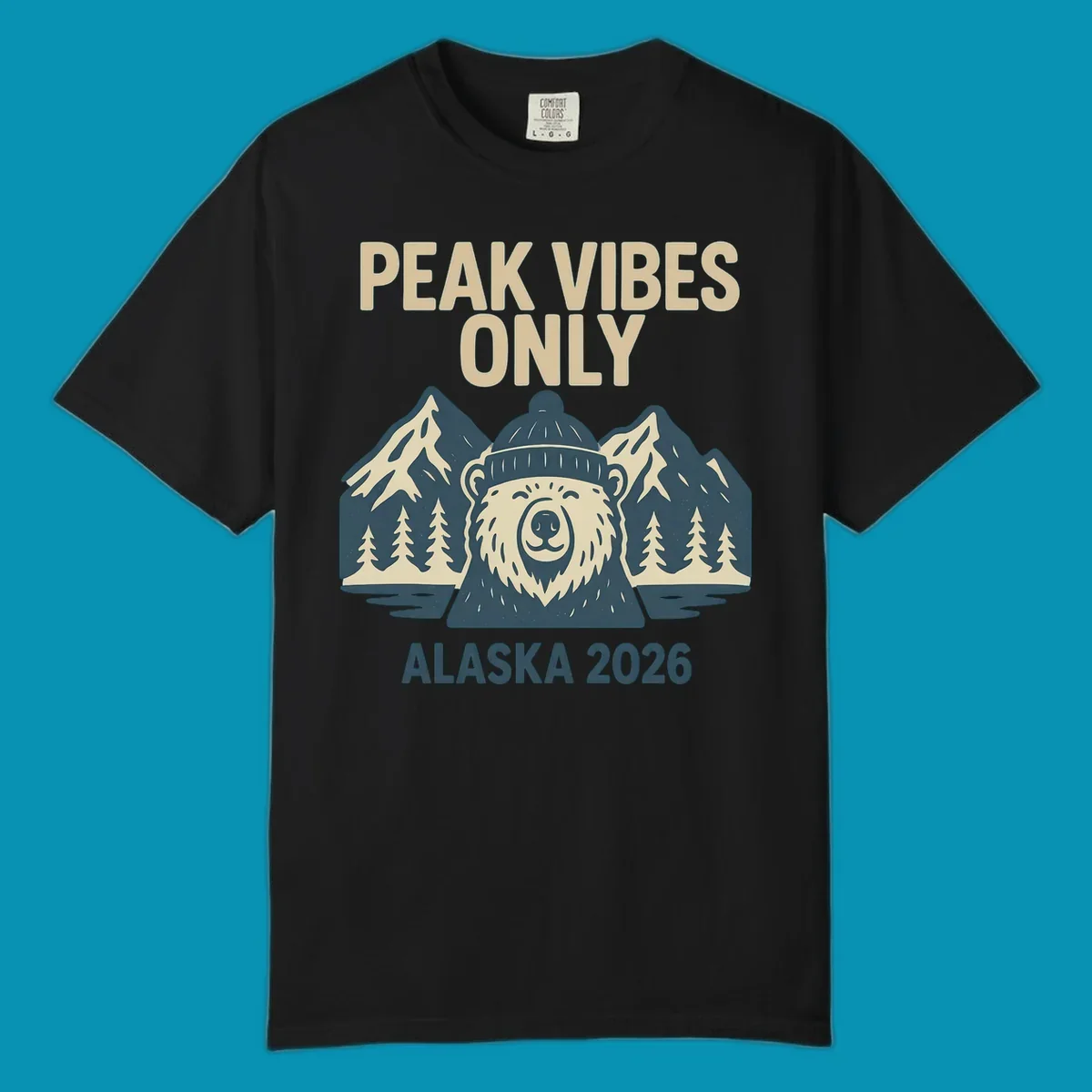 Vintage badge-style illustration of a smiling bear wearing a beanie with snow-capped mountains, evergreen trees, and water, text reads Peak Vibes Only Alaska 2026