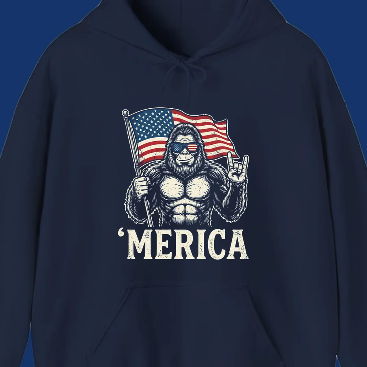 A muscular Bigfoot with American flag sunglasses holds a flag and makes a rock on sign, with "'MERICA" text, for a patriotic Bigfoot 'Merica tee.