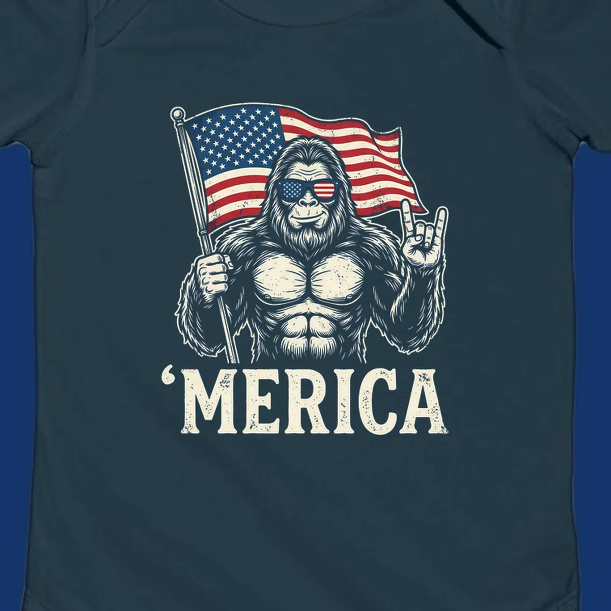 A muscular Bigfoot with American flag sunglasses holds a flag and makes a rock on sign, with "'MERICA" text, for a patriotic Bigfoot 'Merica tee.