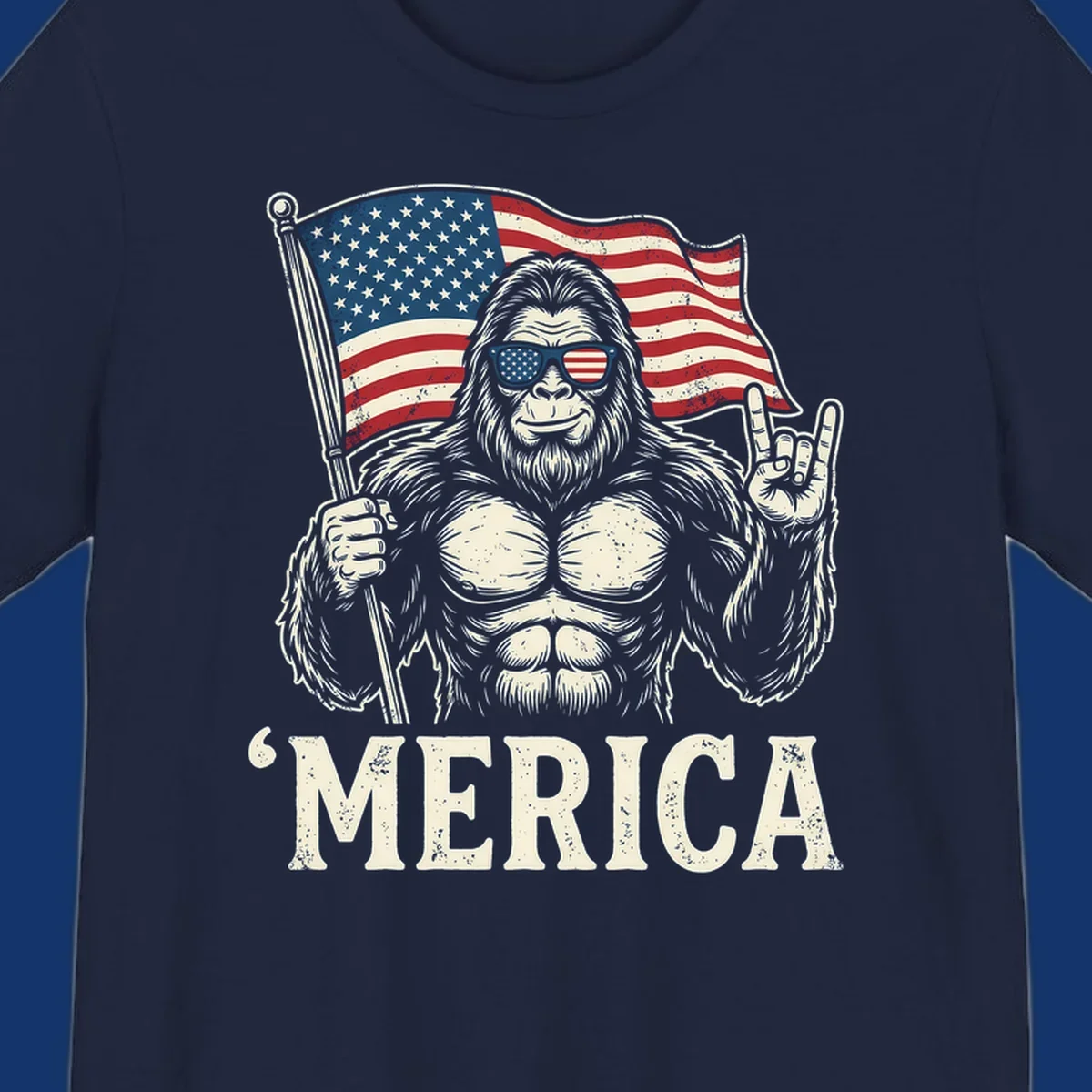 A muscular Bigfoot with American flag sunglasses holds a flag and makes a rock on sign, with "'MERICA" text, for a patriotic Bigfoot 'Merica tee.