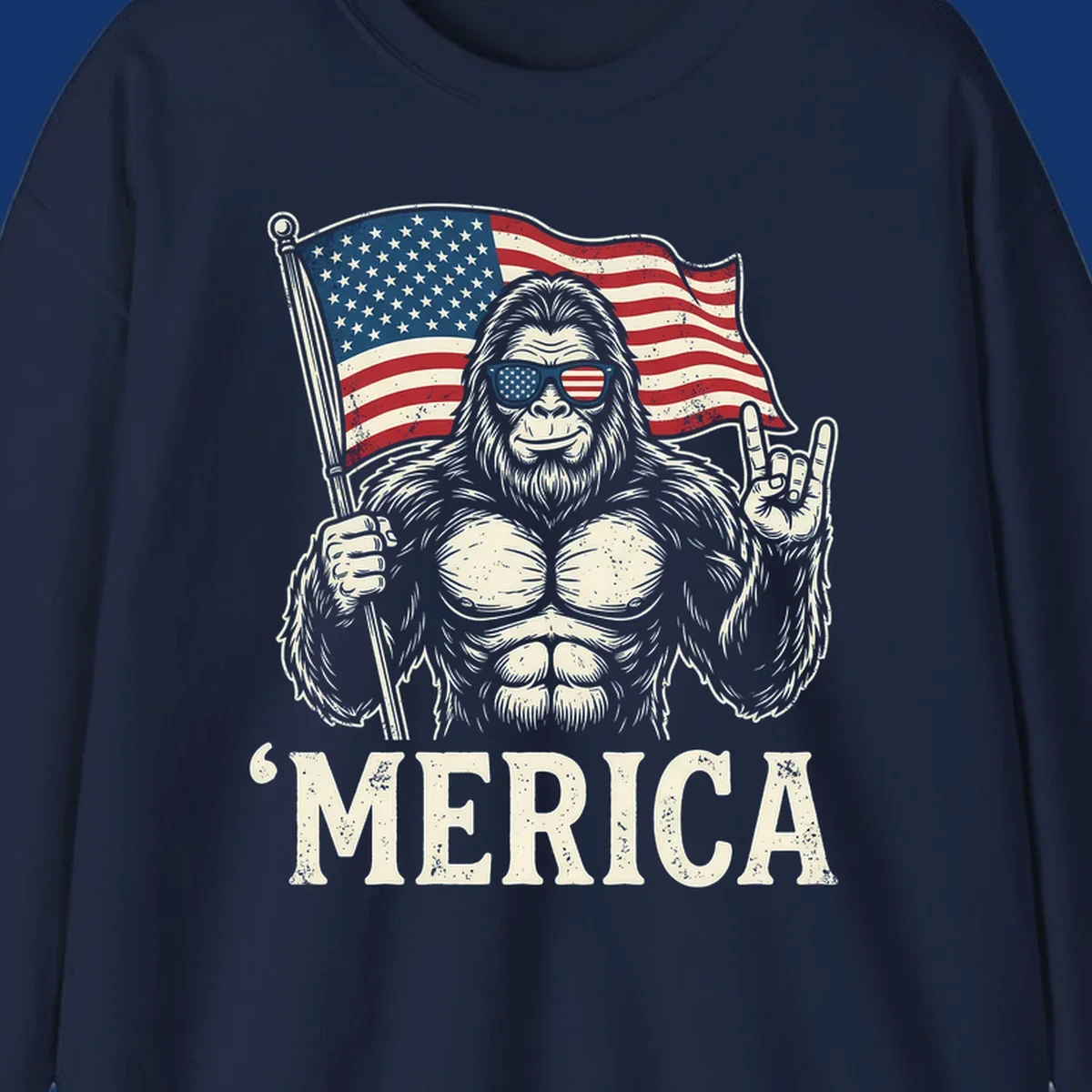 A muscular Bigfoot with American flag sunglasses holds a flag and makes a rock on sign, with "'MERICA" text, for a patriotic Bigfoot 'Merica tee.