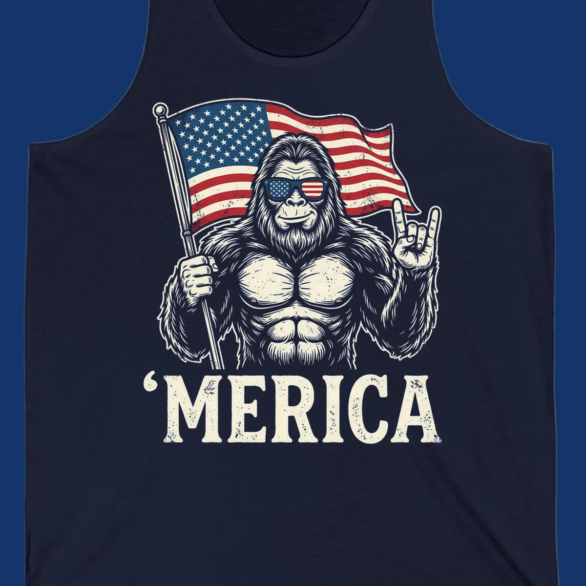 A muscular Bigfoot with American flag sunglasses holds a flag and makes a rock on sign, with "'MERICA" text, for a patriotic Bigfoot 'Merica tee.