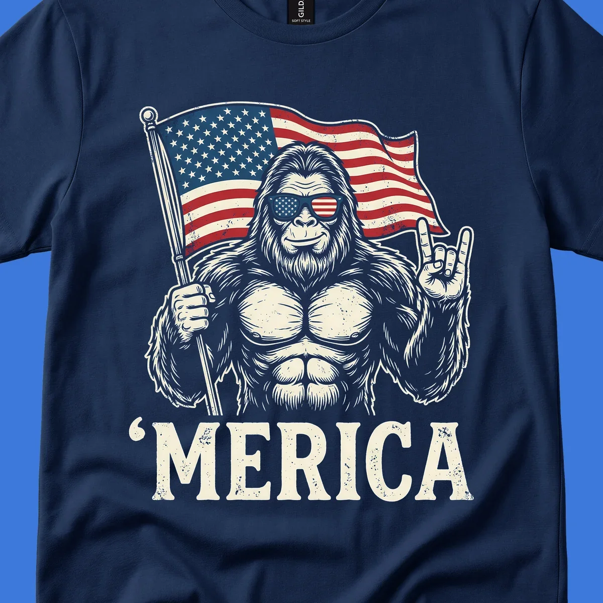 A muscular Bigfoot with American flag sunglasses holds a flag and makes a rock on sign, with "'MERICA" text, for a patriotic Bigfoot 'Merica tee.