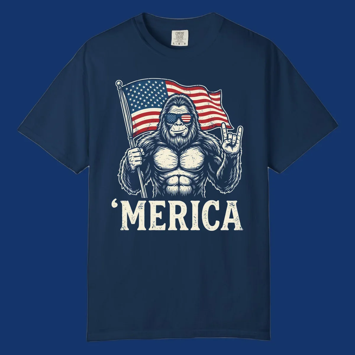 A muscular Bigfoot with American flag sunglasses holds a flag and makes a rock on sign, with "'MERICA" text, for a patriotic Bigfoot 'Merica tee.