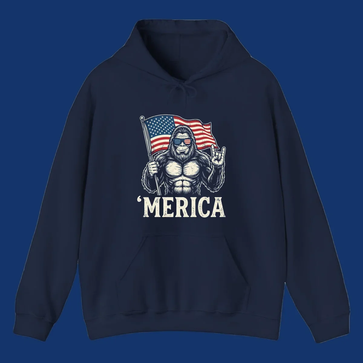 A muscular Bigfoot with American flag sunglasses holds a flag and makes a rock on sign, with "'MERICA" text, for a patriotic Bigfoot 'Merica tee.