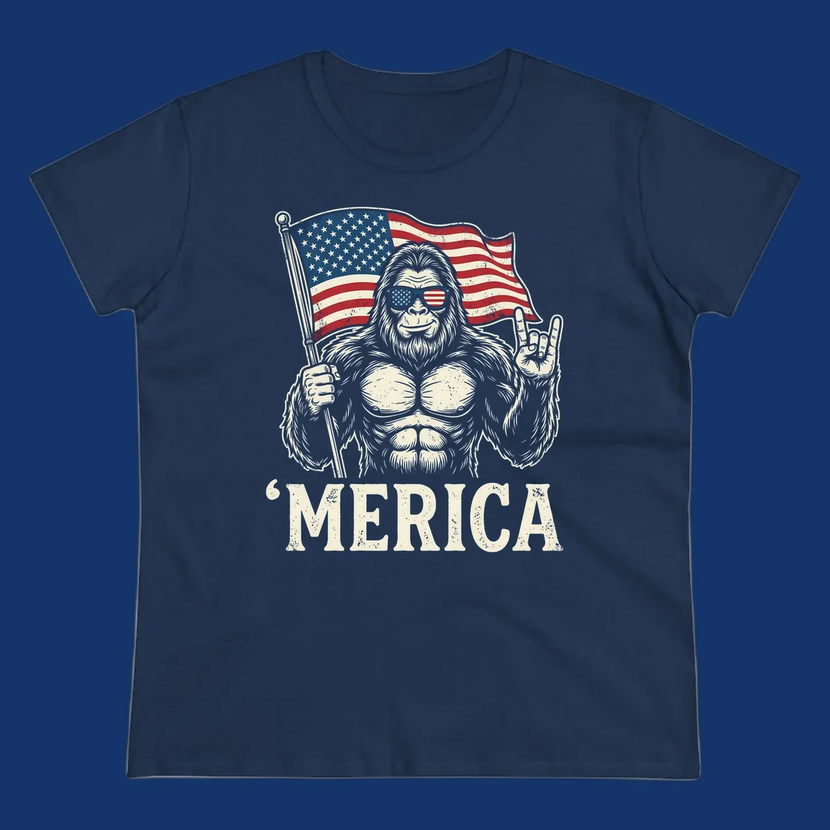 A muscular Bigfoot with American flag sunglasses holds a flag and makes a rock on sign, with "'MERICA" text, for a patriotic Bigfoot 'Merica tee.