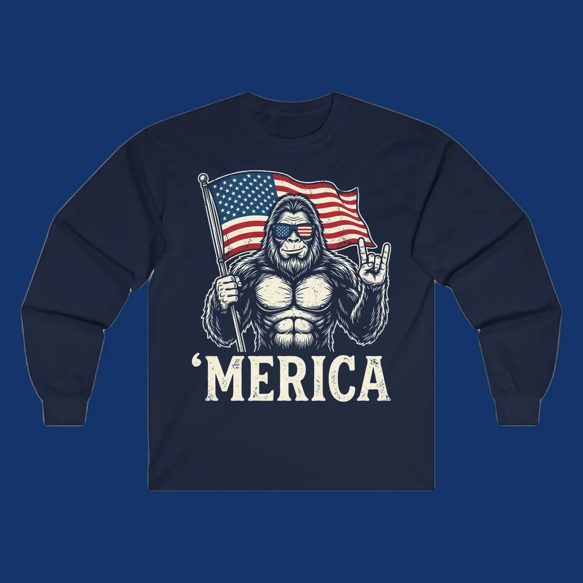 A muscular Bigfoot with American flag sunglasses holds a flag and makes a rock on sign, with "'MERICA" text, for a patriotic Bigfoot 'Merica tee.
