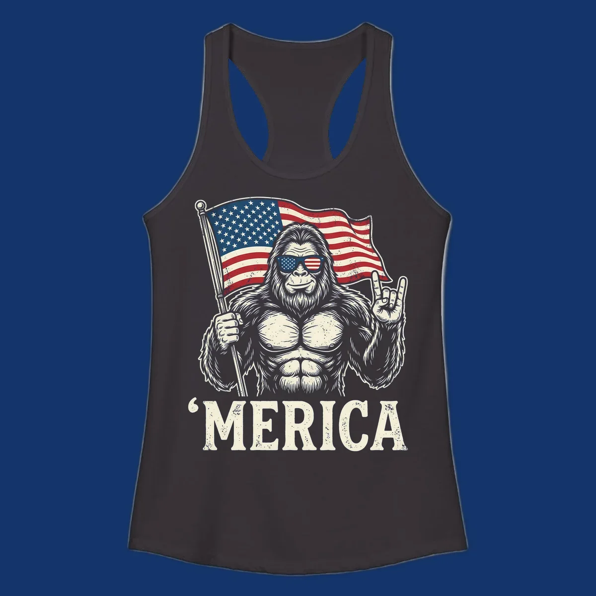 A muscular Bigfoot with American flag sunglasses holds a flag and makes a rock on sign, with "'MERICA" text, for a patriotic Bigfoot 'Merica tee.