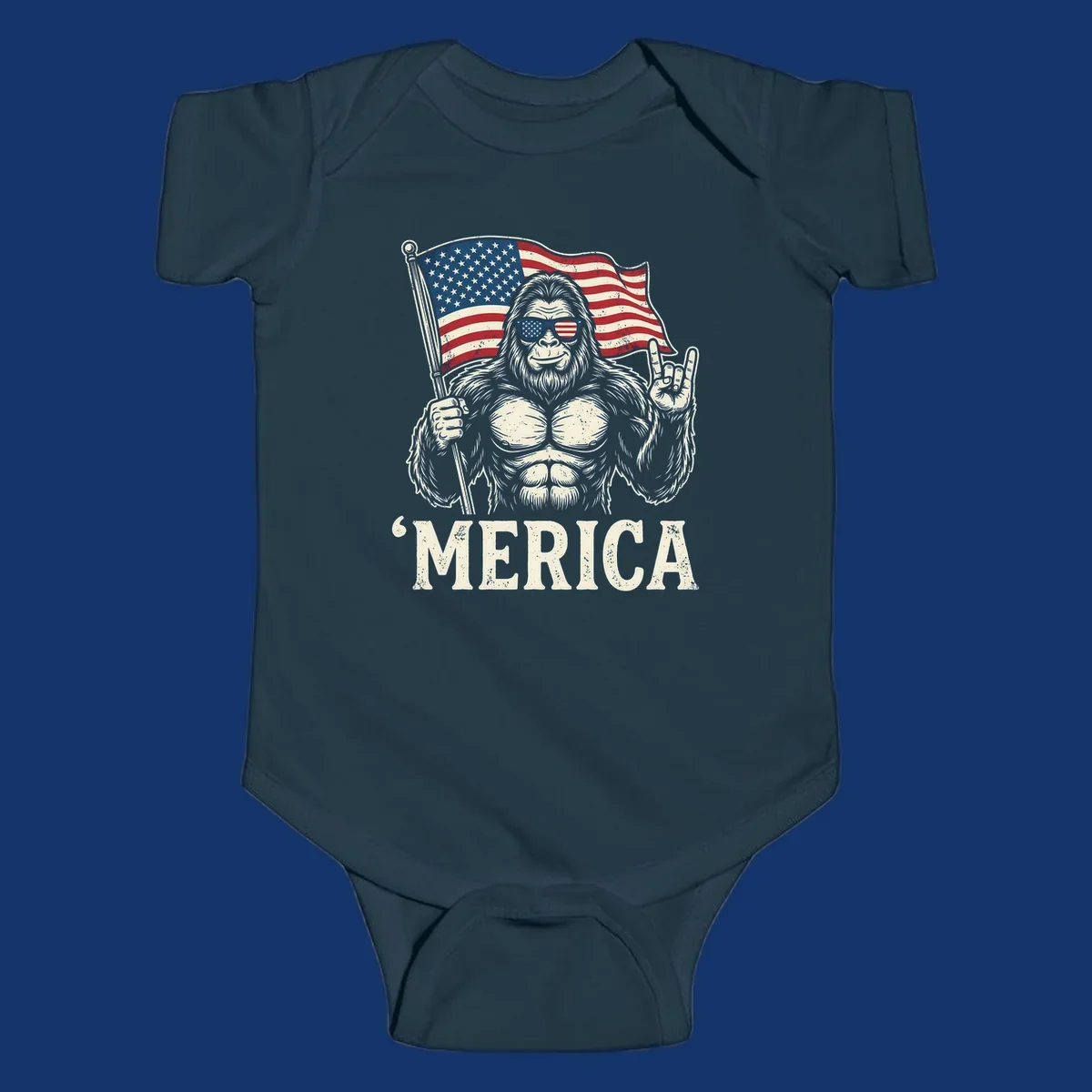 A muscular Bigfoot with American flag sunglasses holds a flag and makes a rock on sign, with "'MERICA" text, for a patriotic Bigfoot 'Merica tee.