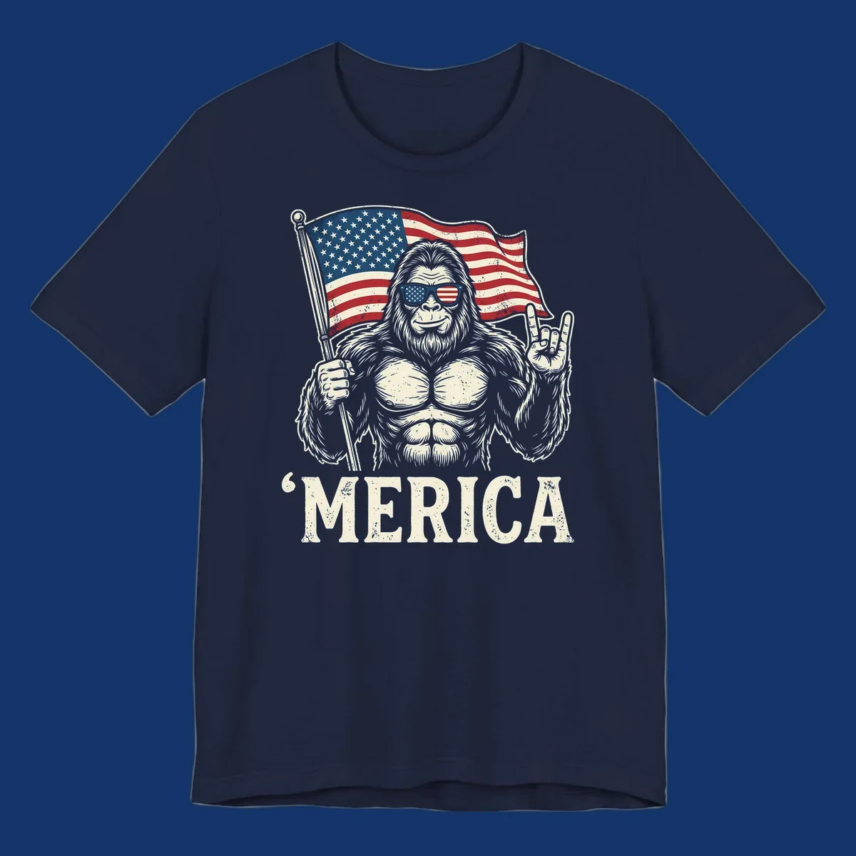 A muscular Bigfoot with American flag sunglasses holds a flag and makes a rock on sign, with "'MERICA" text, for a patriotic Bigfoot 'Merica tee.