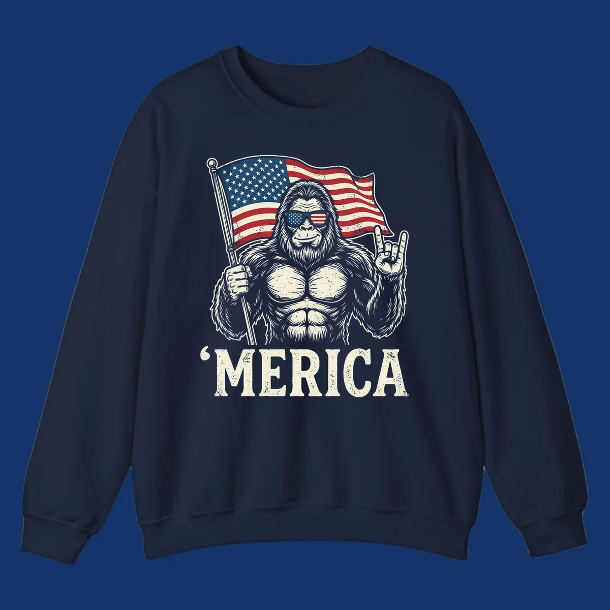 A muscular Bigfoot with American flag sunglasses holds a flag and makes a rock on sign, with "'MERICA" text, for a patriotic Bigfoot 'Merica tee.