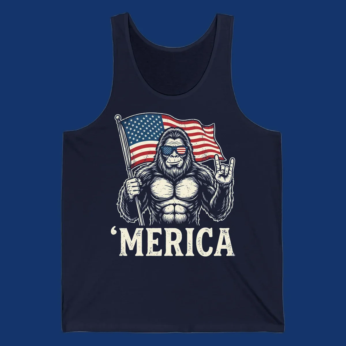 A muscular Bigfoot with American flag sunglasses holds a flag and makes a rock on sign, with "'MERICA" text, for a patriotic Bigfoot 'Merica tee.