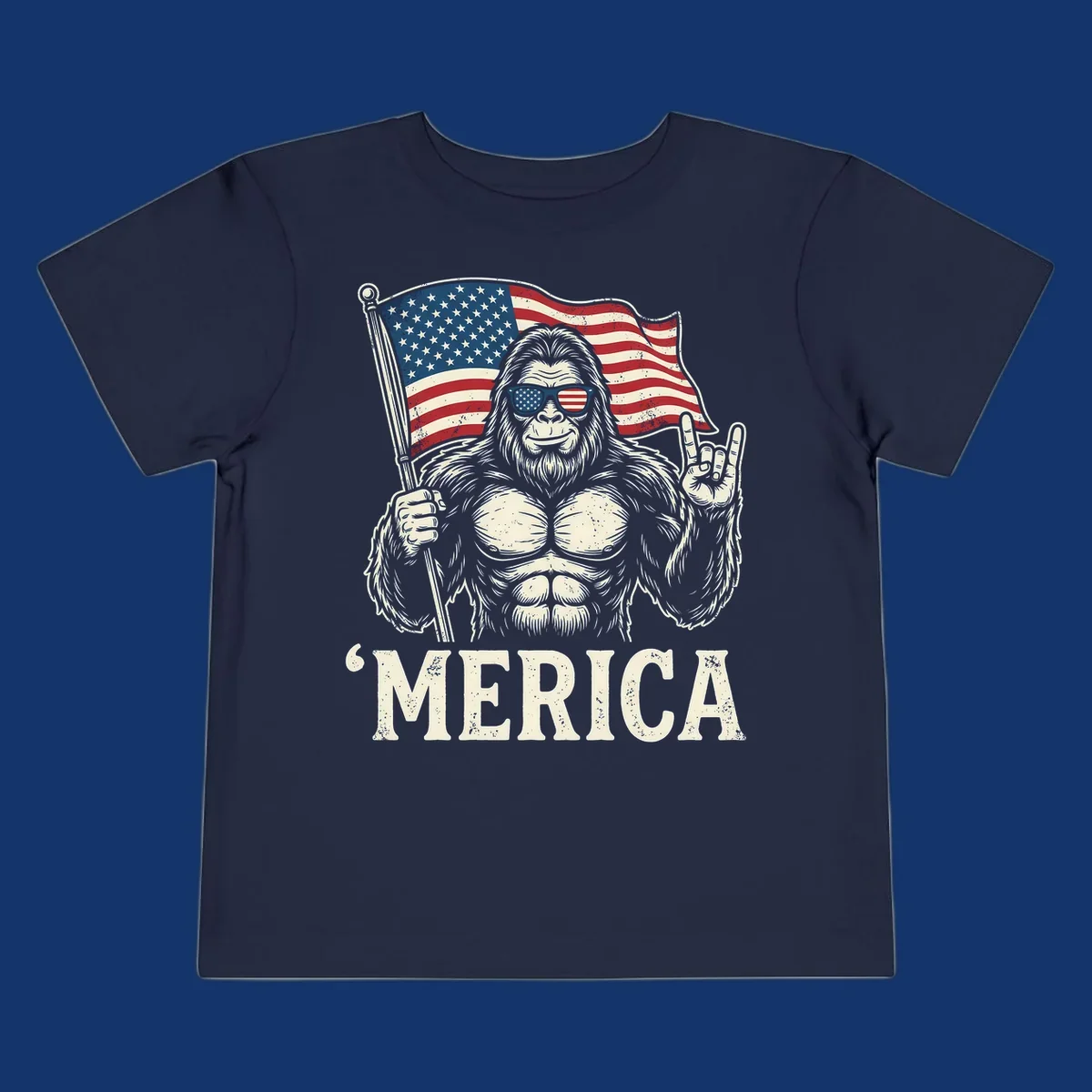 A muscular Bigfoot with American flag sunglasses holds a flag and makes a rock on sign, with "'MERICA" text, for a patriotic Bigfoot 'Merica tee.