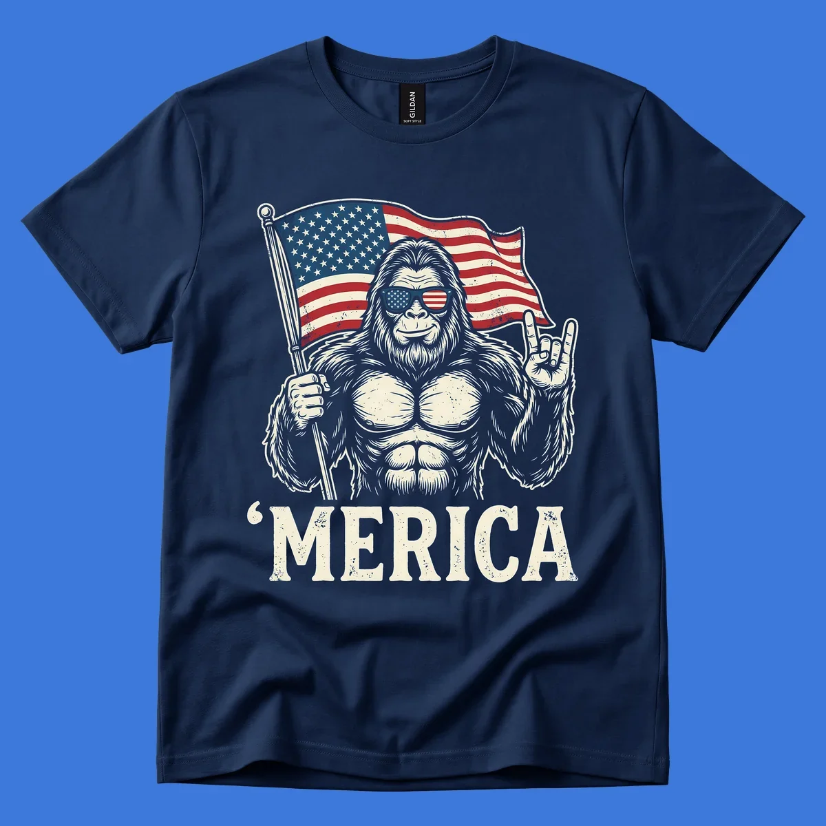 A muscular Bigfoot with American flag sunglasses holds a flag and makes a rock on sign, with "'MERICA" text, for a patriotic Bigfoot 'Merica tee.