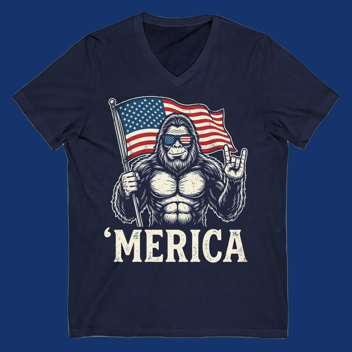 A muscular Bigfoot with American flag sunglasses holds a flag and makes a rock on sign, with "'MERICA" text, for a patriotic Bigfoot 'Merica tee.