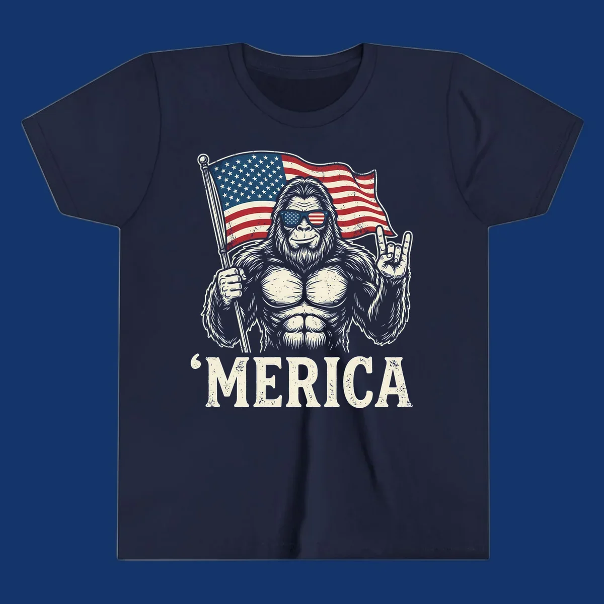A muscular Bigfoot with American flag sunglasses holds a flag and makes a rock on sign, with "'MERICA" text, for a patriotic Bigfoot 'Merica tee.