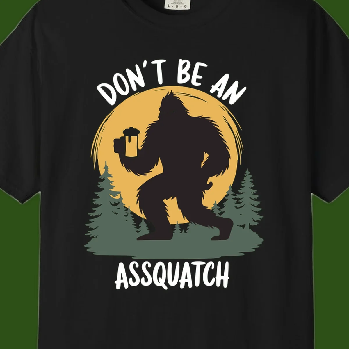 Black silhouette of Bigfoot holding a beer mug under a full moon with pine trees, text reads don't be an assquatch
