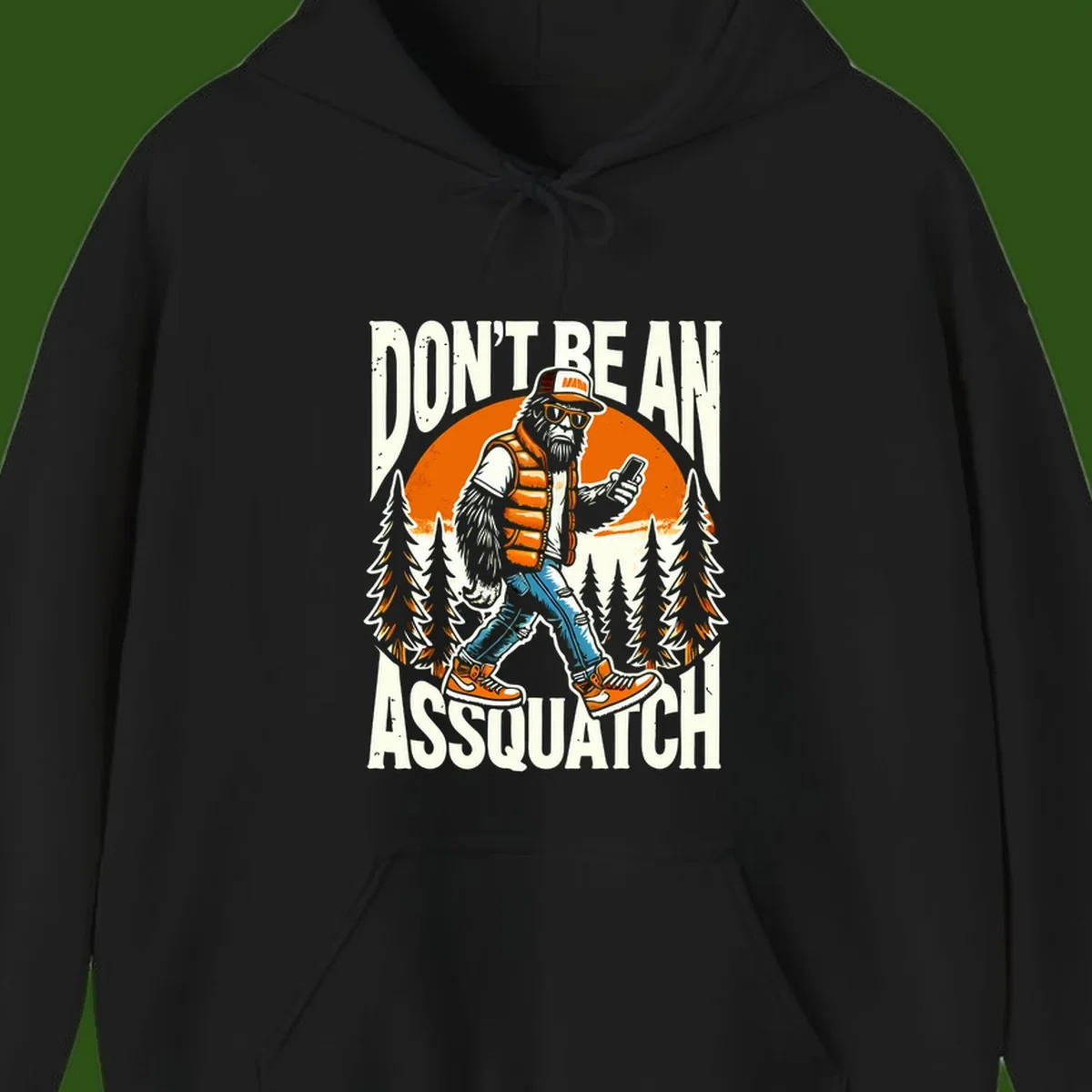 Cartoon sasquatch in hiking gear with backpack and smartphone, 'Don't Be An Assquatch' text, forest and orange sun background.