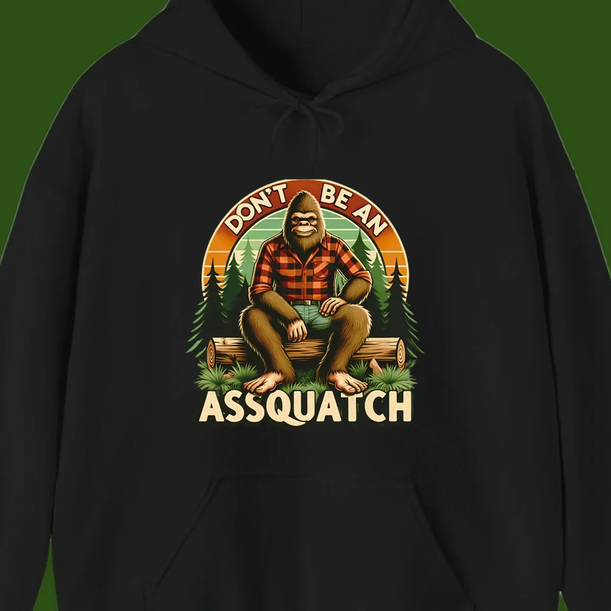 Vintage badge design with friendly Bigfoot in plaid shirt sitting on a log with forest trees, text reads 'Don't Be An Assquatch'