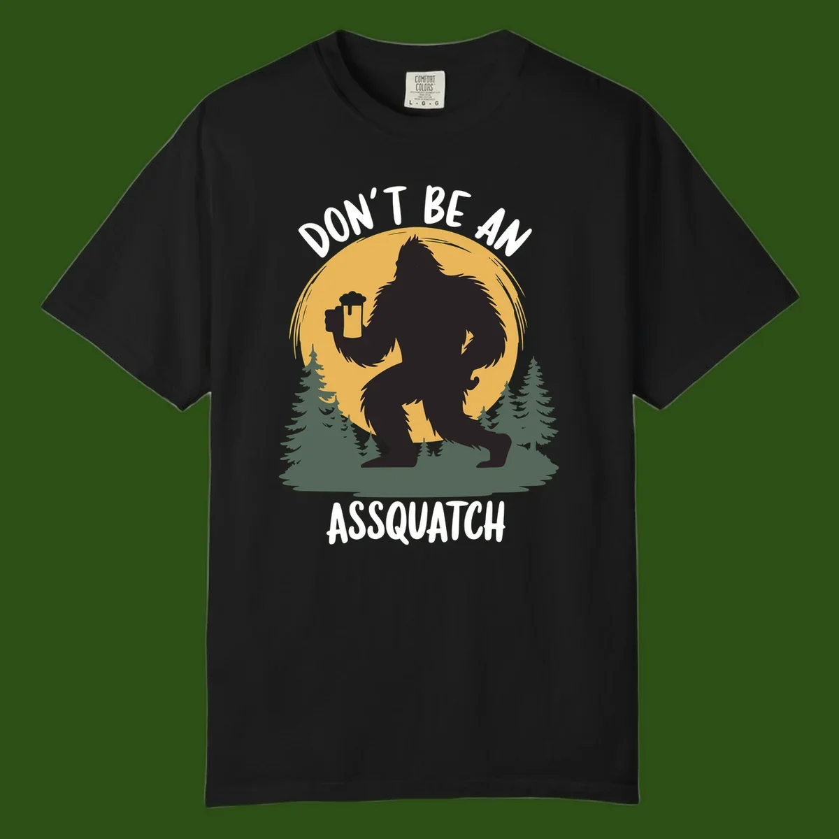 Black silhouette of Bigfoot holding a beer mug under a full moon with pine trees, text reads don't be an assquatch