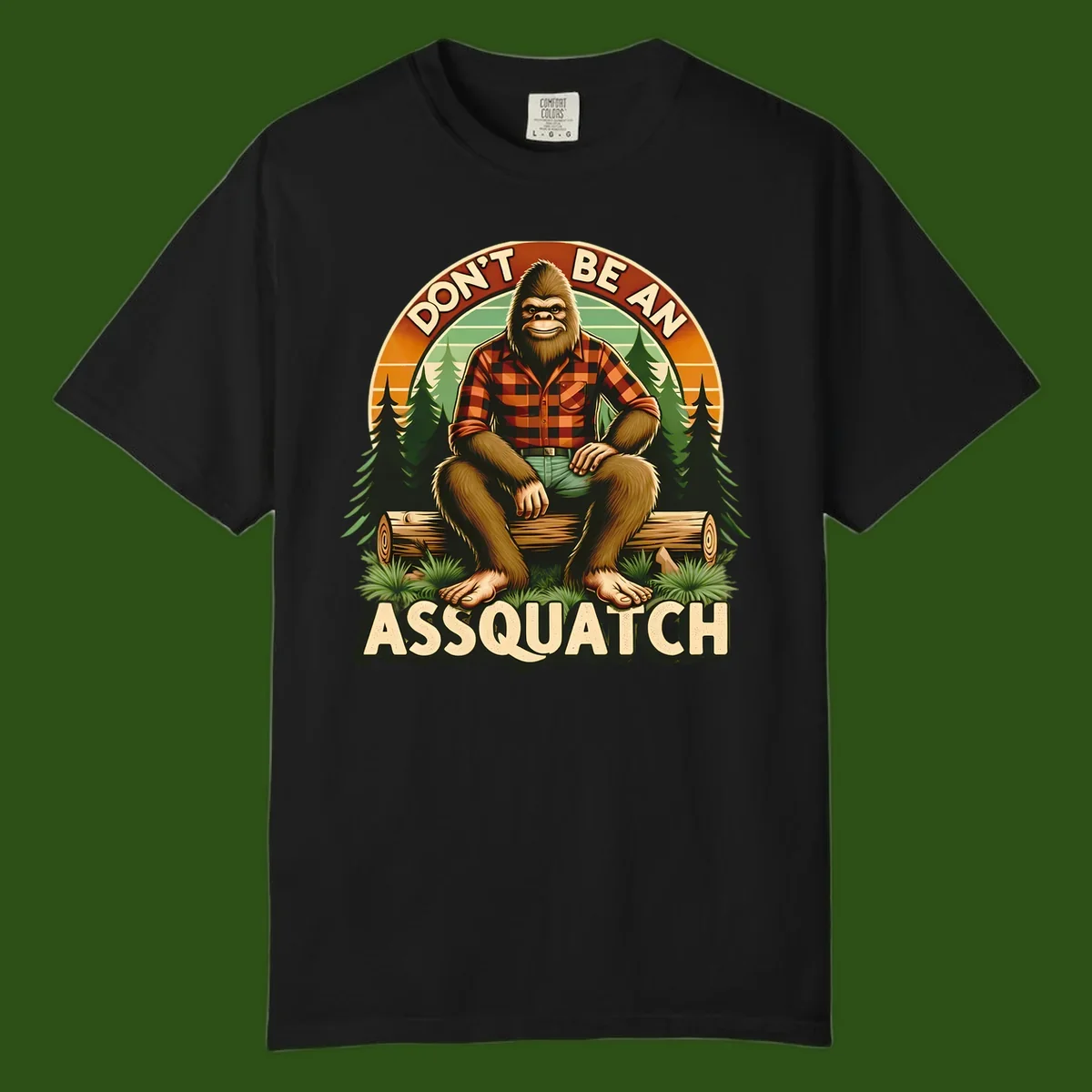 Vintage badge design with friendly Bigfoot in plaid shirt sitting on a log with forest trees, text reads 'Don't Be An Assquatch'