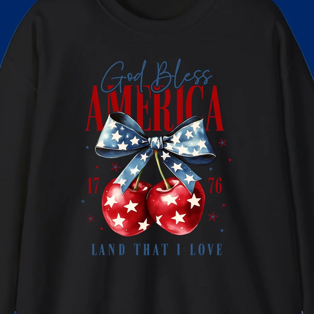 A patriotic design featuring 'God Bless AMERICA' and 'LAND THAT I LOVE' with a blue star-spangled bow, two red star-covered cherries, and the numbers '17' and '76'. god-bless-america-cherries.