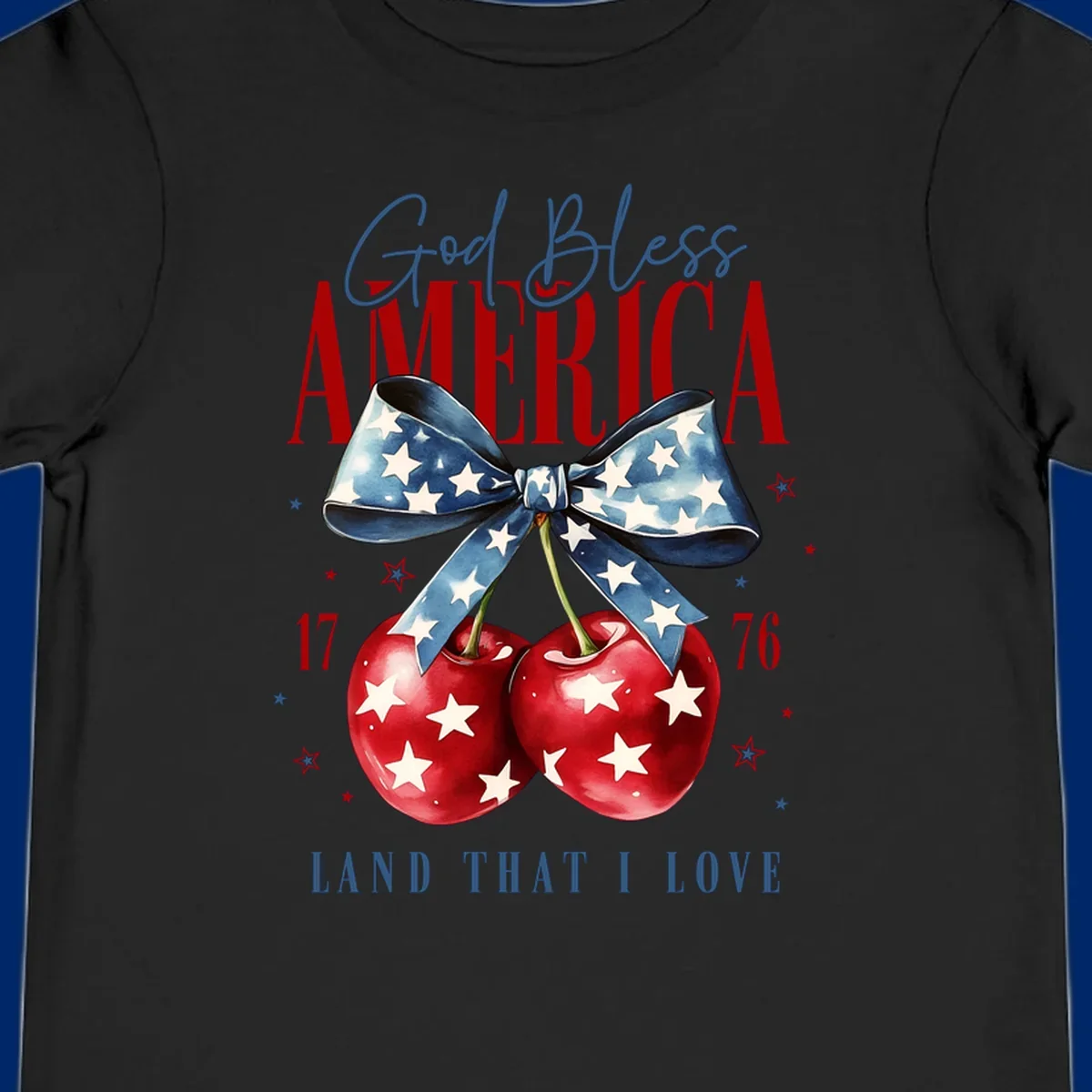 A patriotic design featuring 'God Bless AMERICA' and 'LAND THAT I LOVE' with a blue star-spangled bow, two red star-covered cherries, and the numbers '17' and '76'. god-bless-america-cherries.
