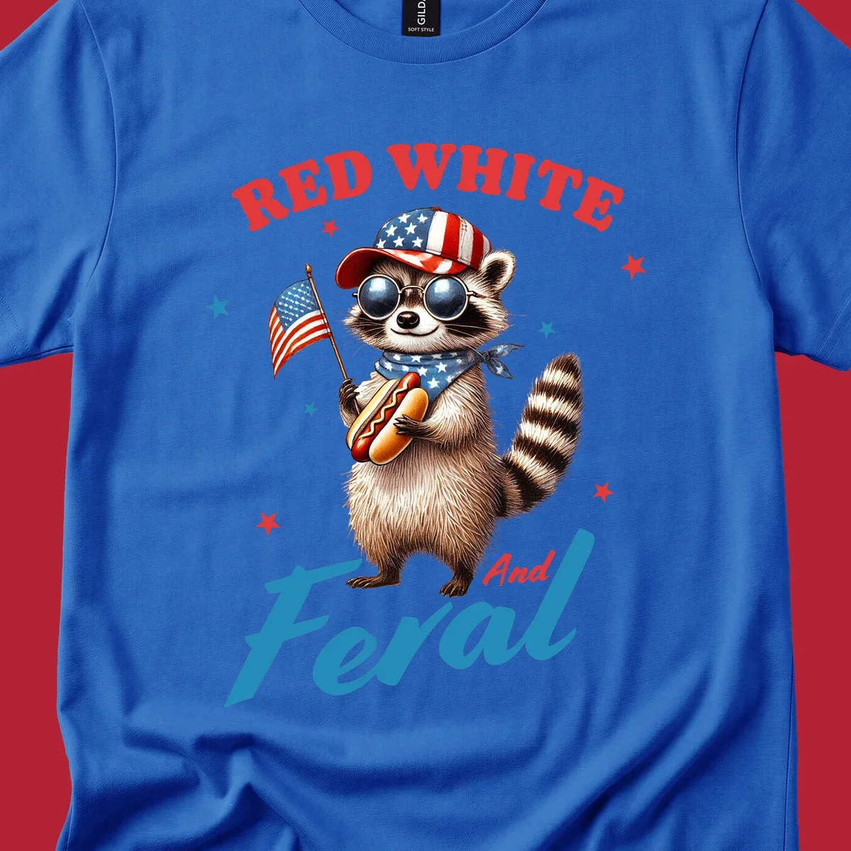 A cartoon raccoon wearing a patriotic hat and bandana, holding an American flag and a hot dog, with 'Red White And Feral' text. This is a red-white-feral-raccoon design.