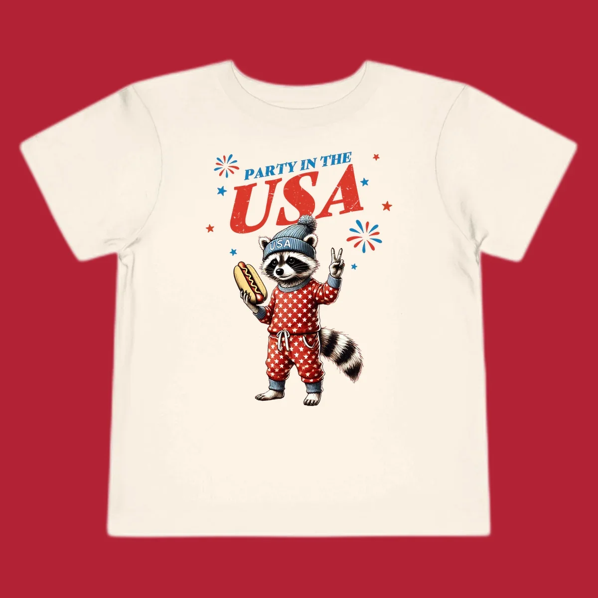 A cartoon raccoon wearing a USA beanie and star-patterned pajamas, holding a hotdog and making a peace sign, with 'PARTY IN THE USA' text and fireworks.