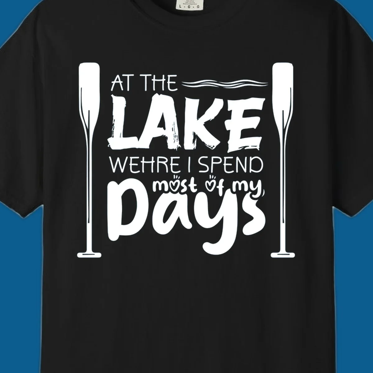 Text design "At The Lake Where I Spend Most Of My Days" with paddles and waves, lake-life-days concept.