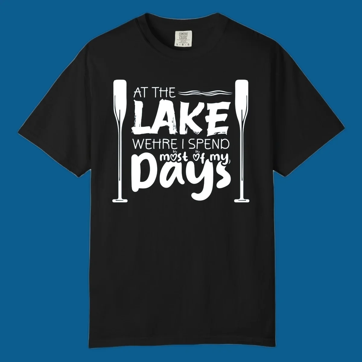 Text design "At The Lake Where I Spend Most Of My Days" with paddles and waves, lake-life-days concept.