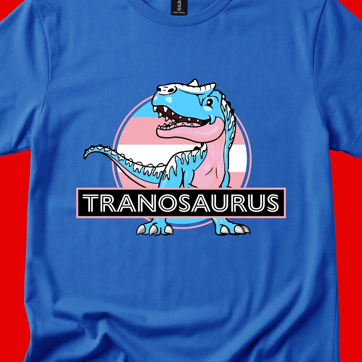 Blue cartoon T-Rex dinosaur with open mouth against pink and white pride flag circular background, labeled Tranosaurus