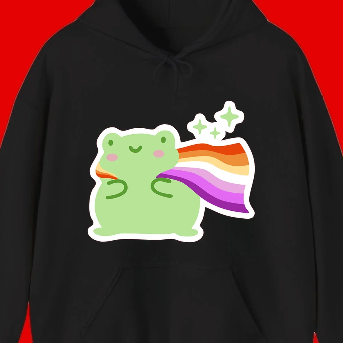 Cute green frog with pink cheeks wearing a rainbow pride cape, surrounded by sparkle stars
