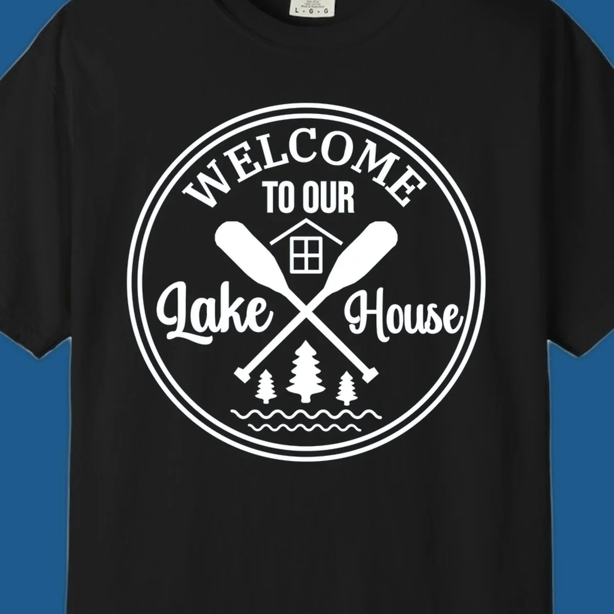 Circular badge design with text 'WELCOME TO OUR Lake House', crossed oars, house, trees, and waves.